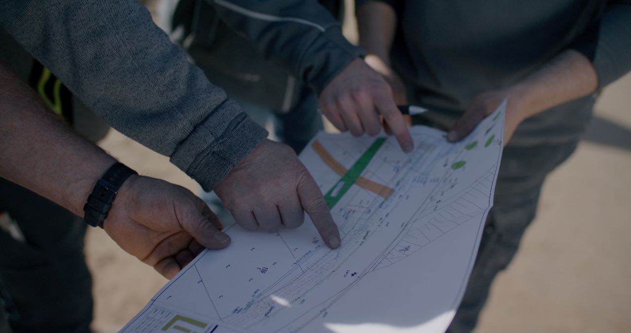 Drei Personen besprechen technische Zeichnungen einer Ausführungsplanung im Freien auf einer Baustelle
