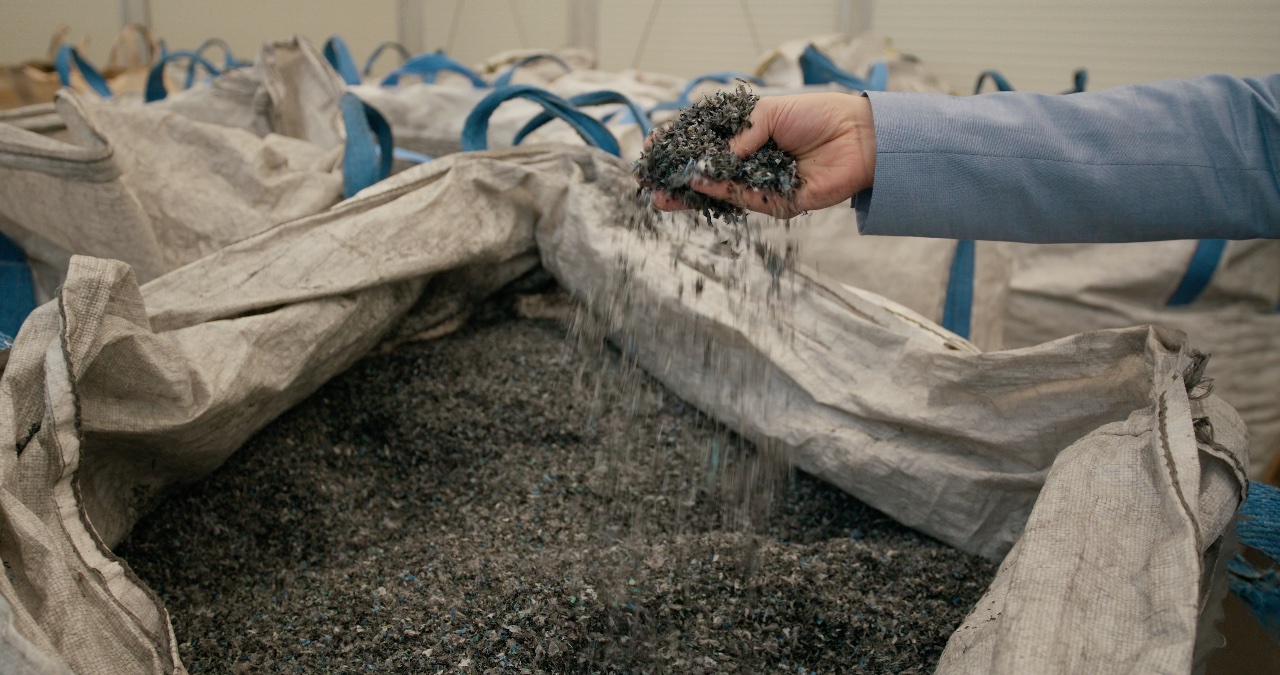 Hand prüft dunkles Kunststoff-Rezyklat in großen Big-Bags innerhalb einer Industriehalle