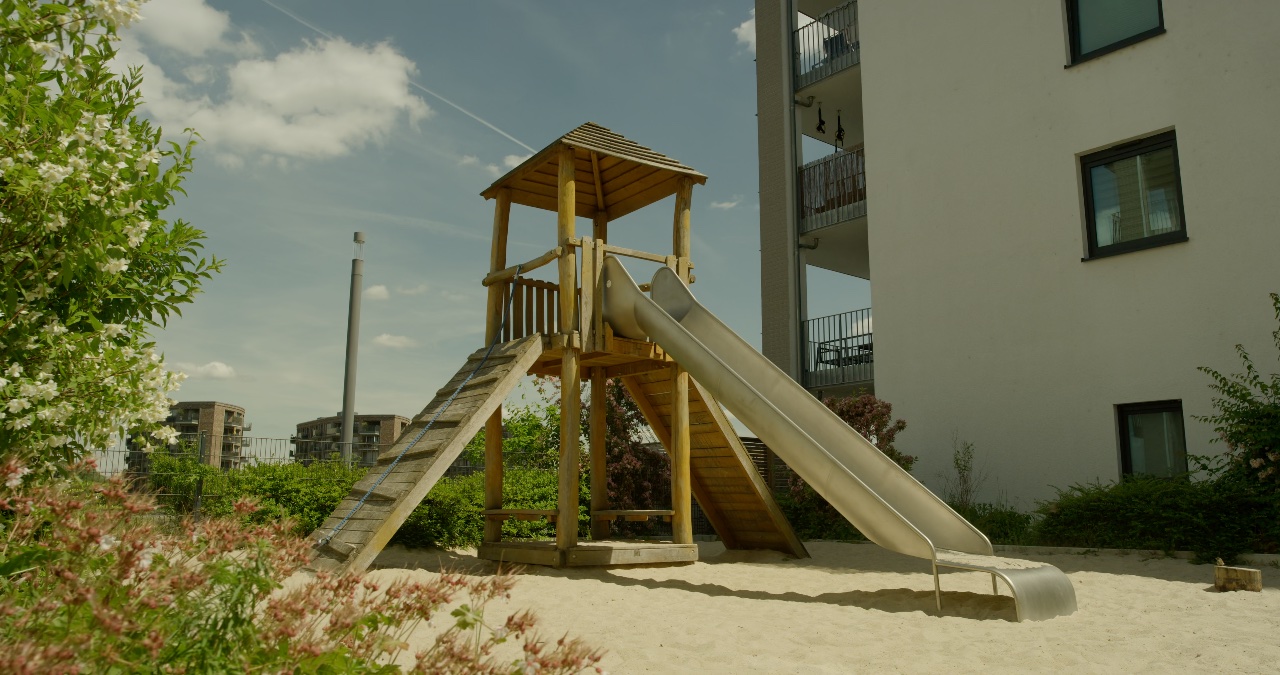 Holzturm mit Rutsche und Klettermöglichkeit auf einem Sandspielplatz vor modernen Wohngebäuden an einem sonnigen Tag