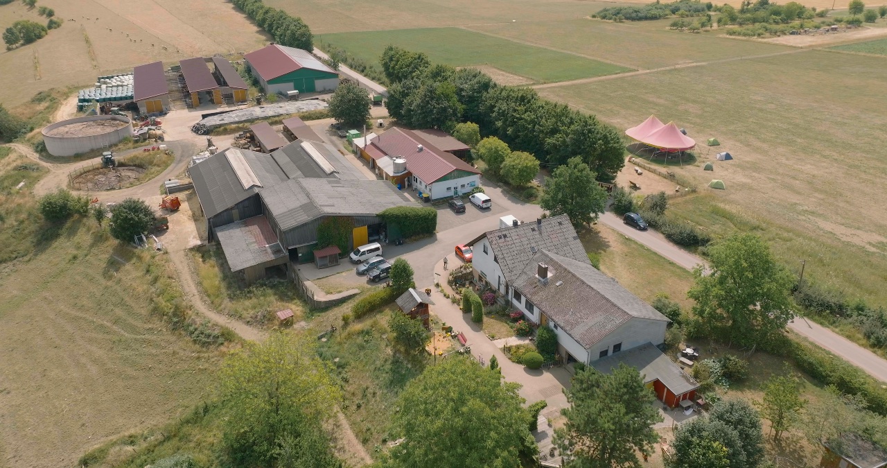 Luftaufnahme eines weitläufigen Bauernhofs mit Stallungen, Wohnhäusern und angrenzenden Feldern