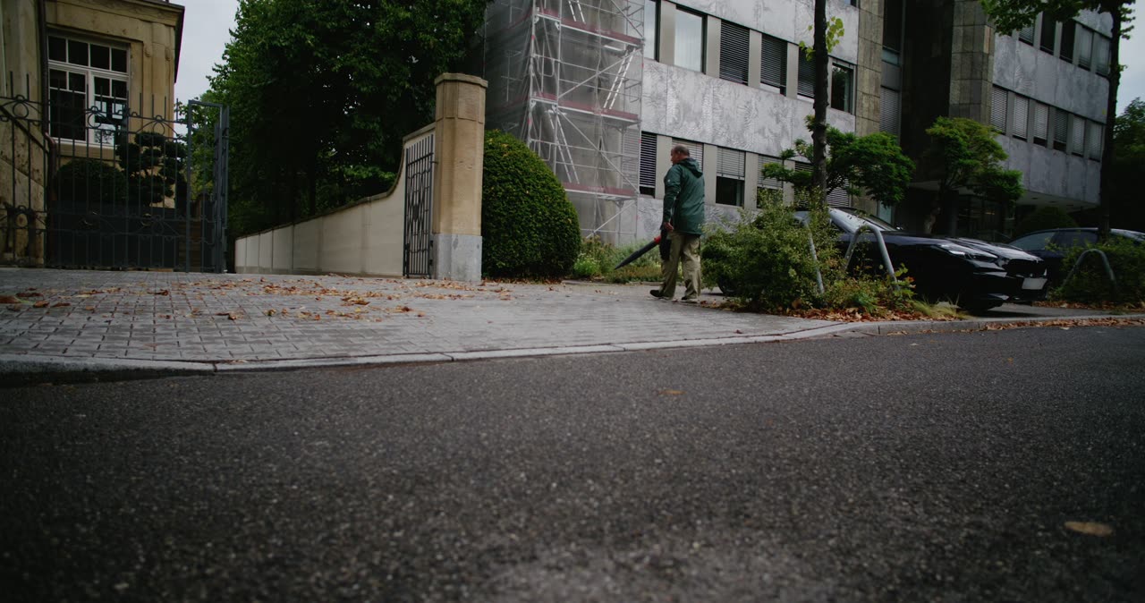 Mann mit Laubbläser beim Reinigen von einem gepflasterten Gehweg neben einer Straße im städtischen Raum