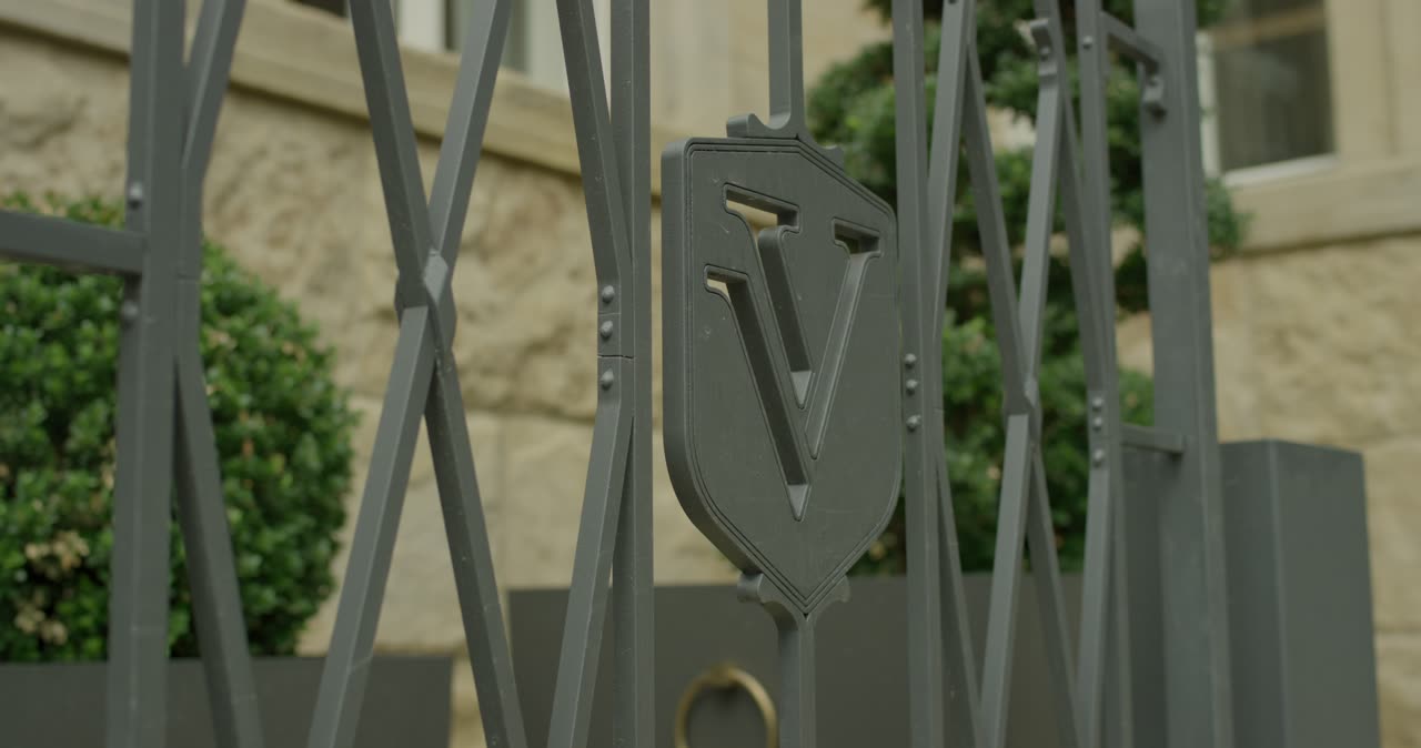 Nahaufnahme von einem grauen Metalltor mit einem wappenförmigen V-Logo vor einer Sandsteinfassadetage